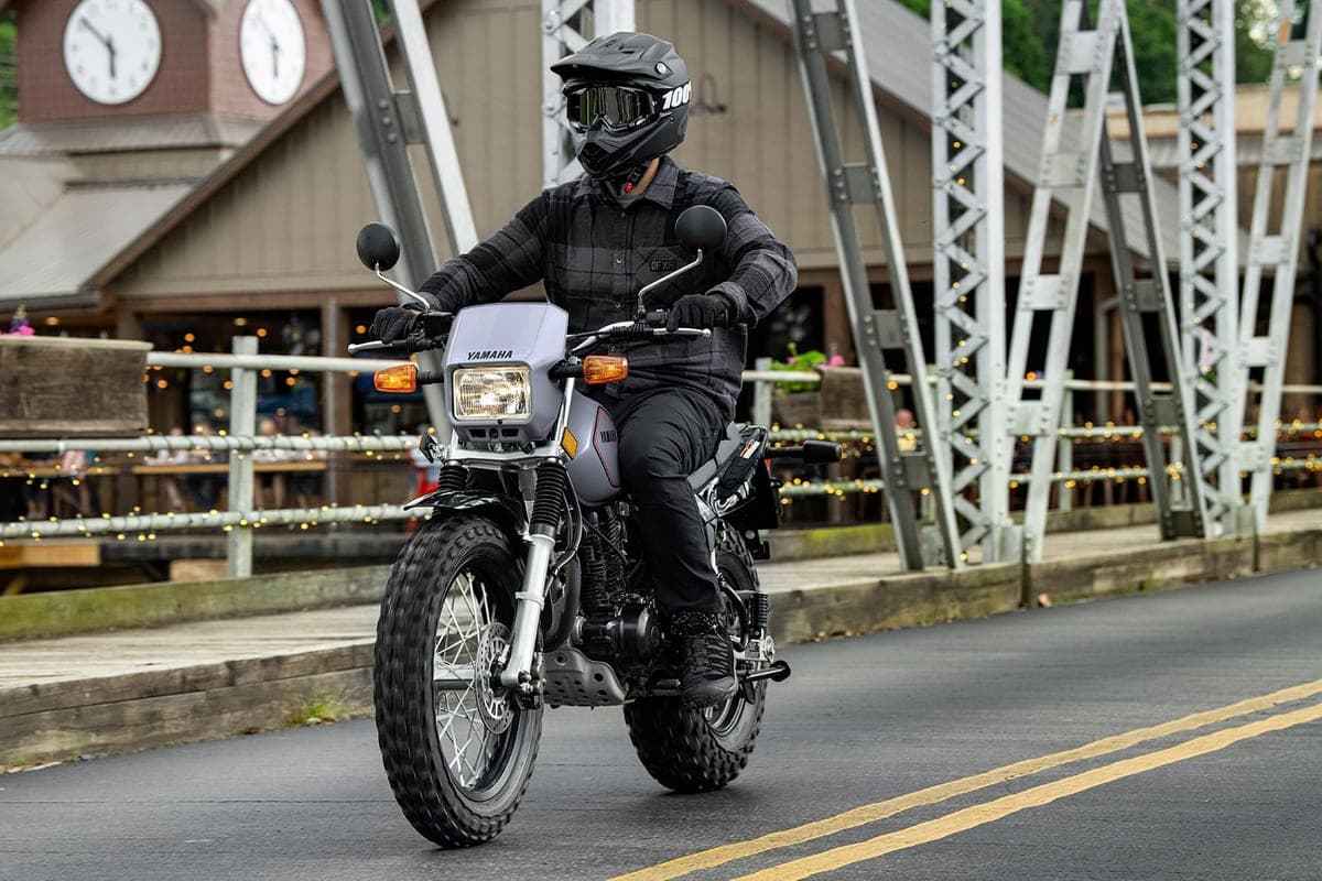 Rider in dark plaid jacket and full-face helmet riding a silver Yamaha TW200 across a small-town bridge with string lights and a clock tower behind