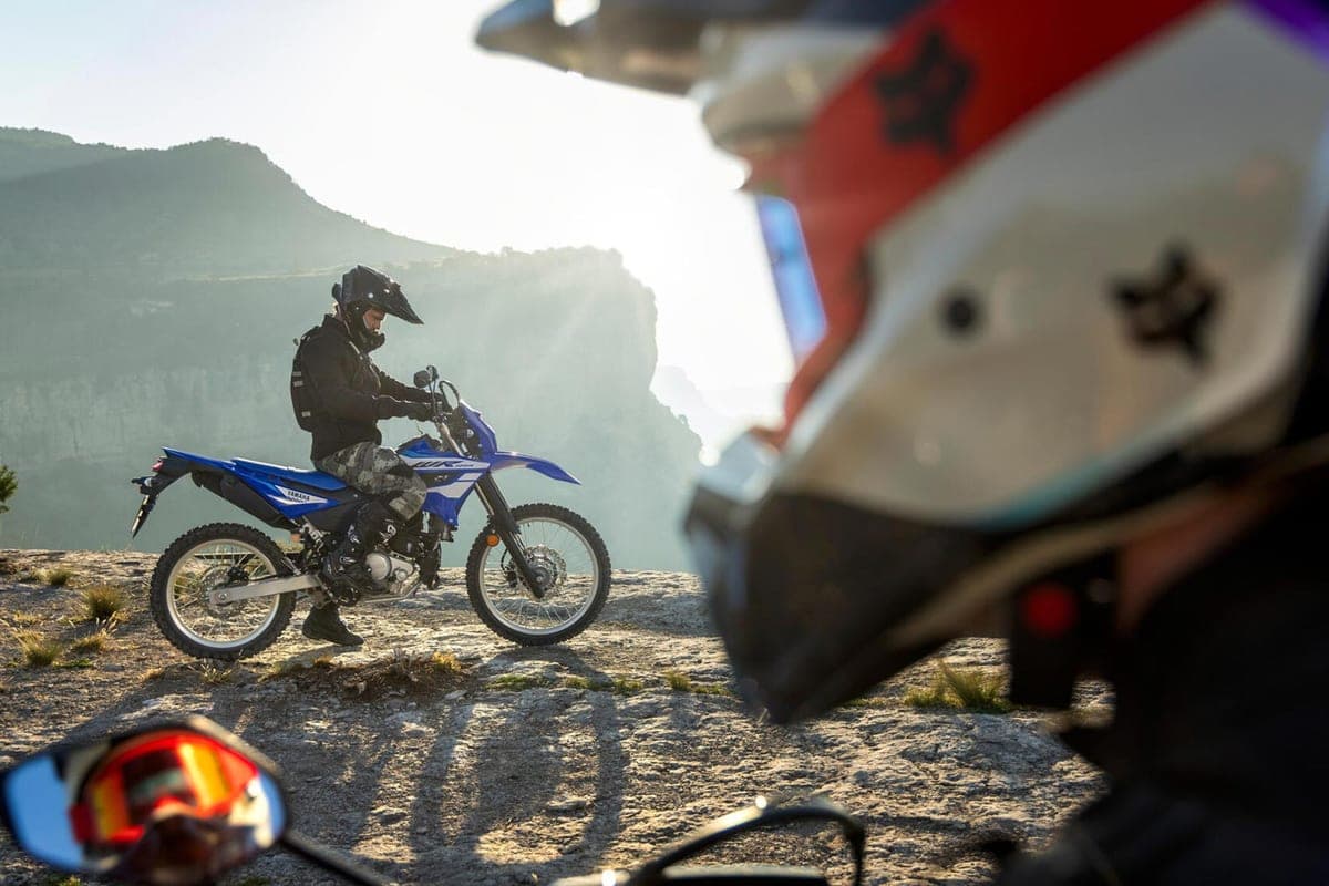 Rider in dark gear pausing a blue Yamaha WR125R on a rocky overlook at sunset, with helmets and goggles visible in the foreground