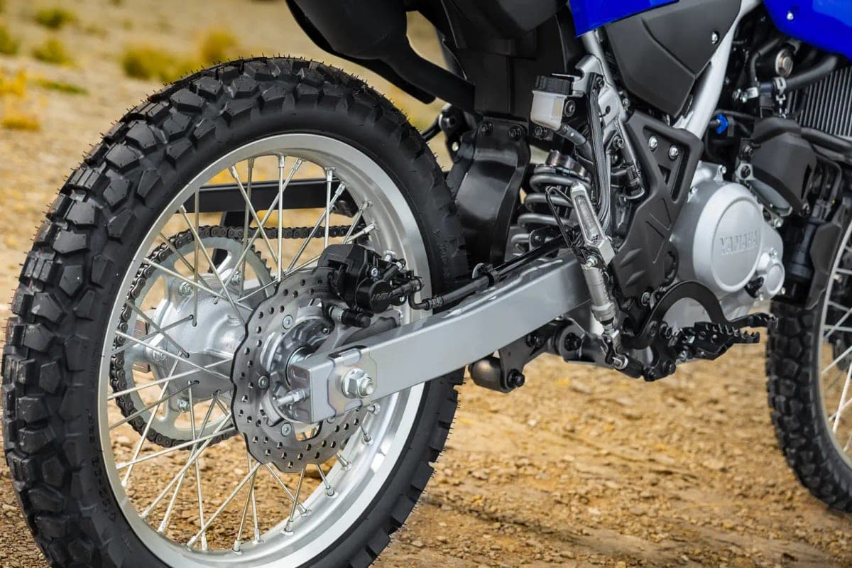 Close-up of the WR125R's rear wheel, chain, disc brake and shock linkage against a dirt trail background