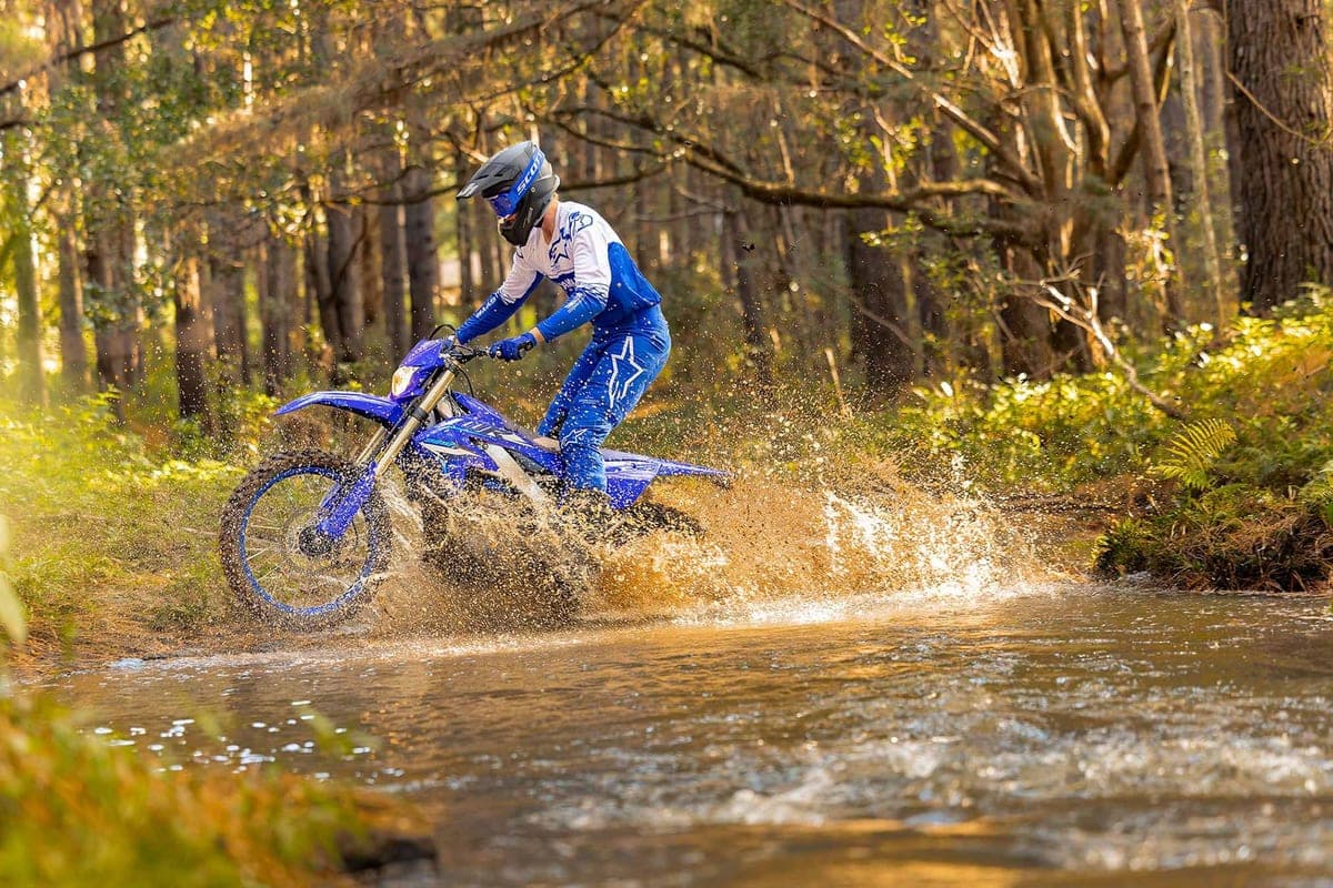 Rider in blue and white Alpinestars gear blasting a blue Yamaha WR250F through a shallow forest stream, throwing up a wide spray of muddy water