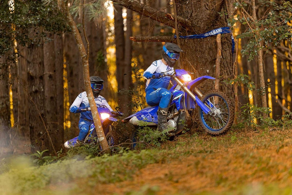 Two riders on blue Yamaha WR-series enduros pausing on a wooded trail at dusk with headlights lit, a Yamaha racing banner tied to a nearby tree