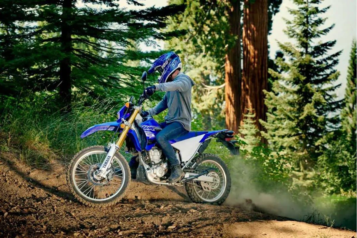 Rider in blue helmet and grey jacket climbing a dusty dirt trail on a Yamaha WR250R, kicking up a cloud of dust through evergreen forest