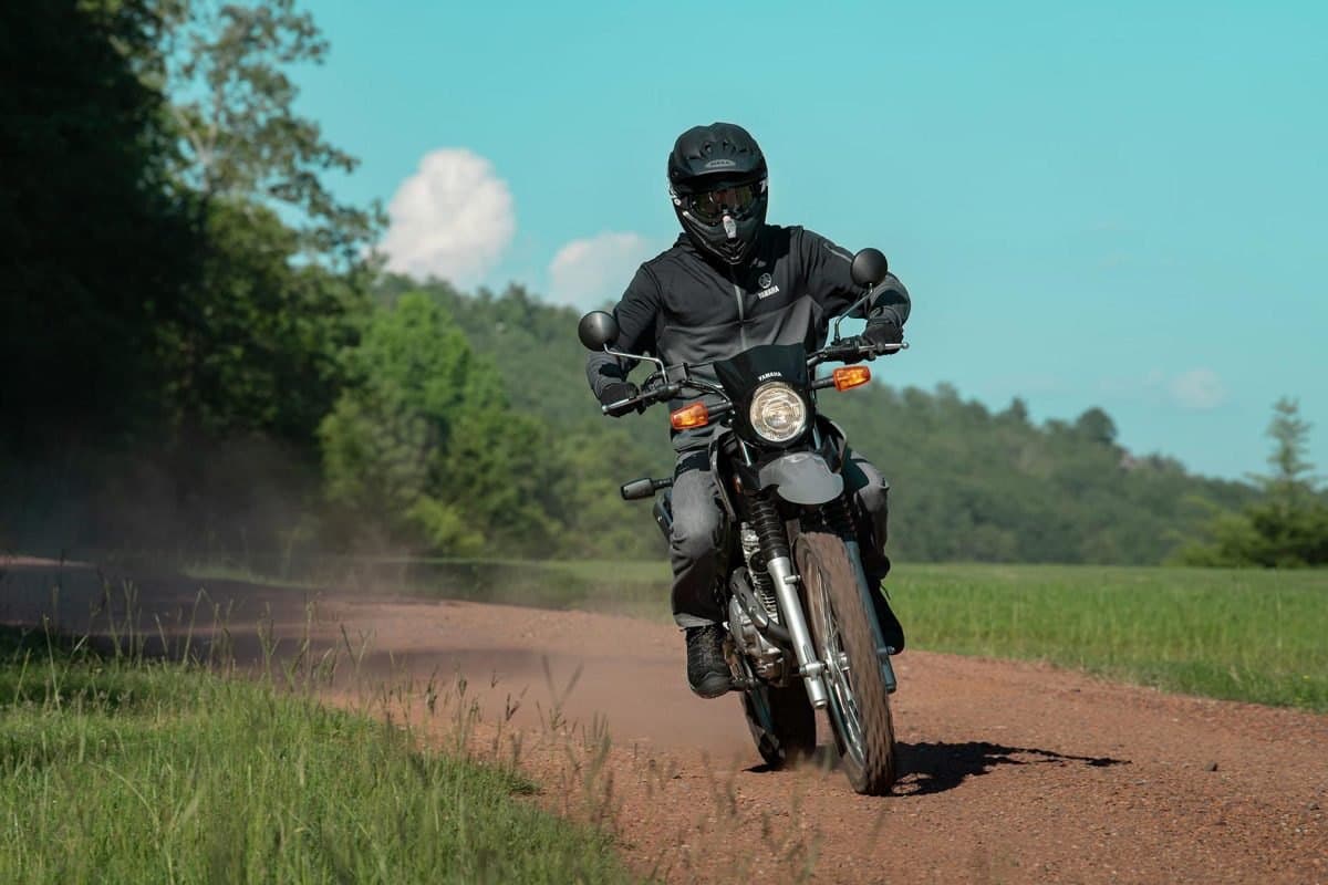 Rider in black gear riding a grey Yamaha XT250 down a dusty red-dirt road, kicking up a cloud of dust with green fields on either side