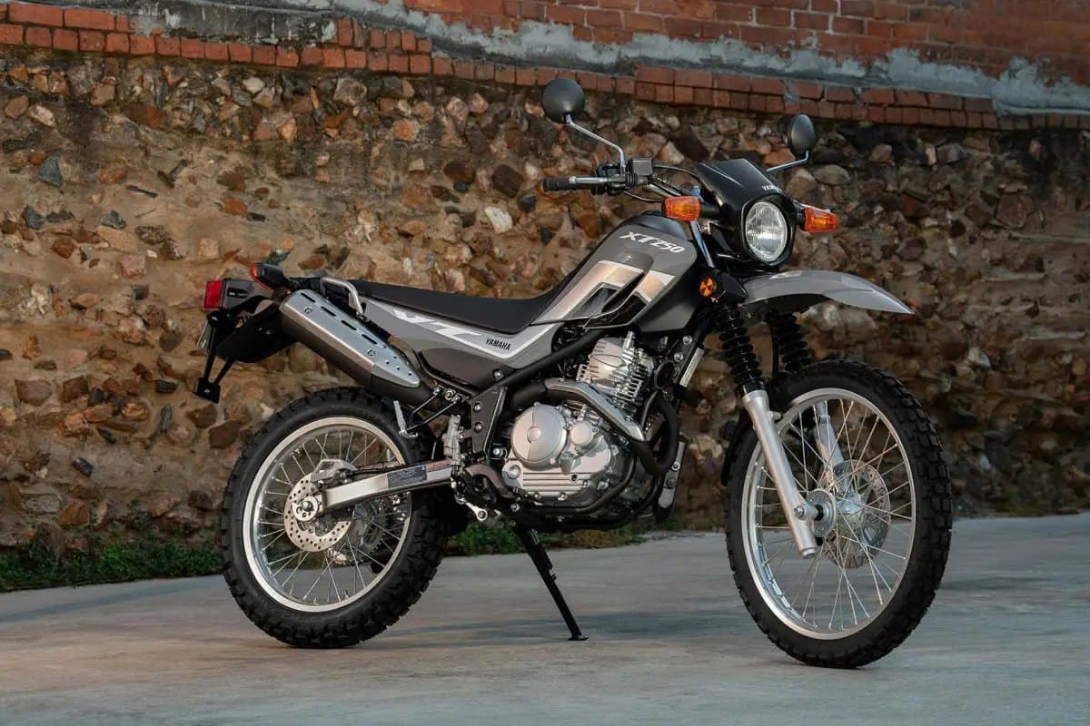 2025 Yamaha XT250 front three-quarter studio view against a stone wall backdrop, showing the round headlight, long-travel forks and skid-equipped exhaust
