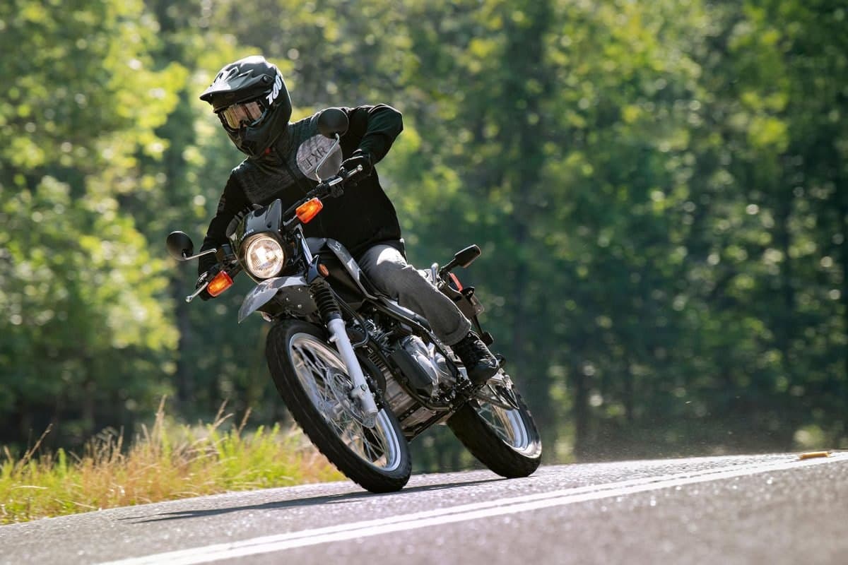 Rider in dark gear leaning a Yamaha XT250 through a paved corner, front wheel cutting a tight line with a sunlit forest blurred behind