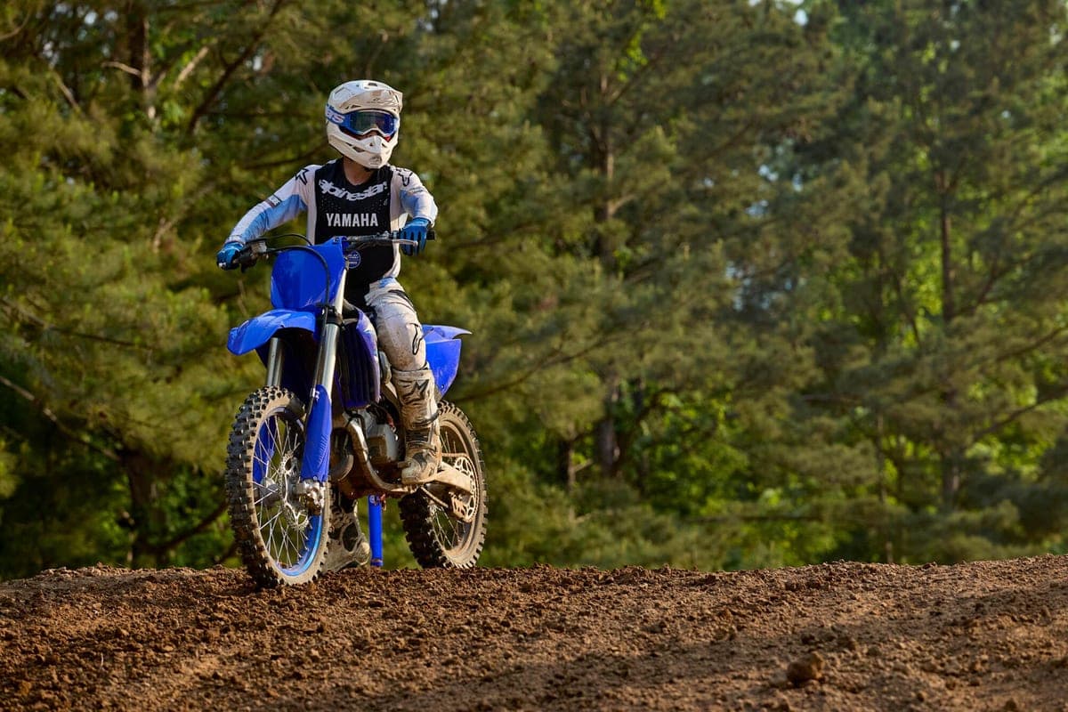 Rider in white and black Yamaha gear cresting a dirt berm on a blue YZ125 two-stroke, muddy boots and forest backdrop behind