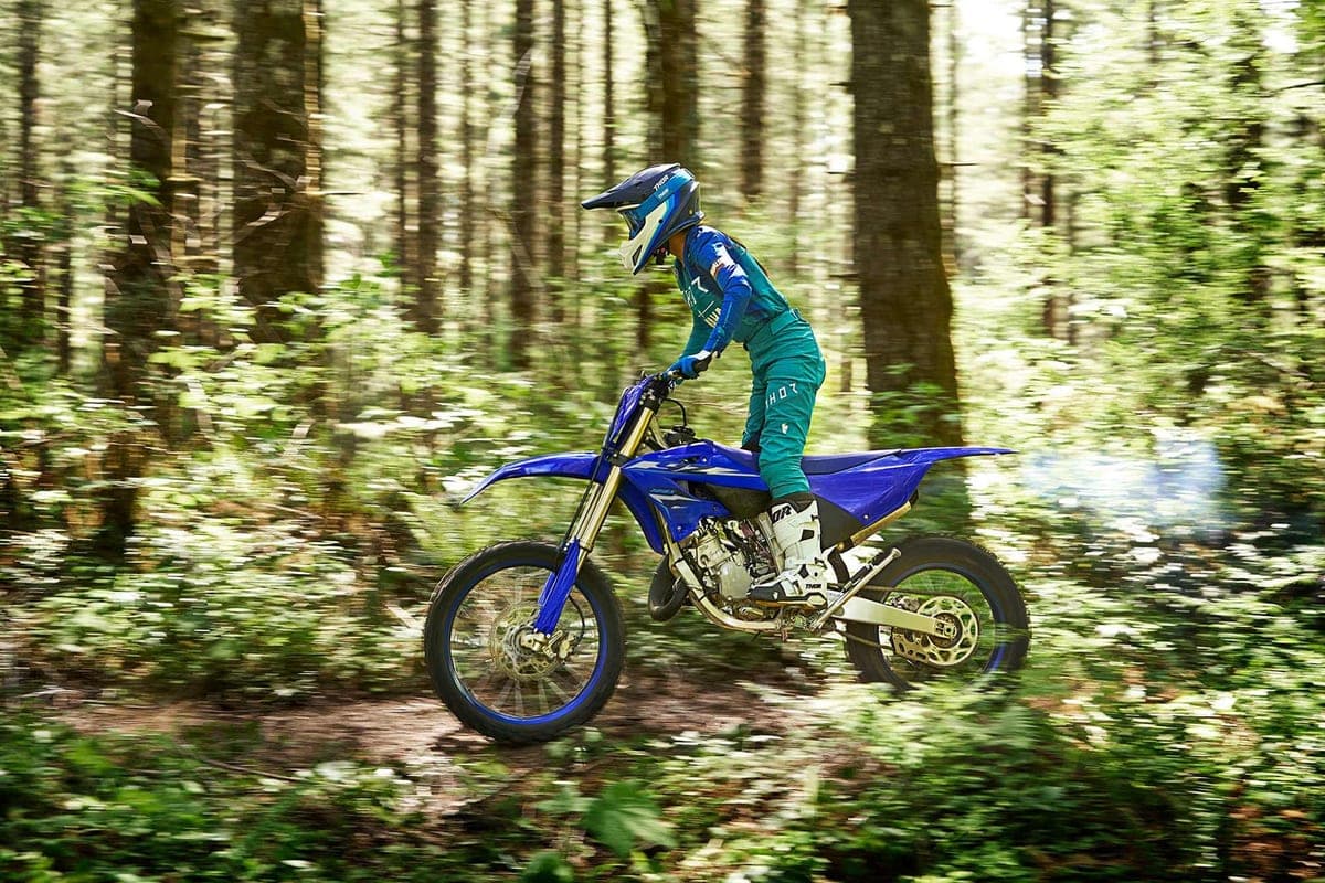 Rider in teal gear and a helmet catching air on a blue Yamaha YZ125X along a shaded singletrack trail through tall forest
