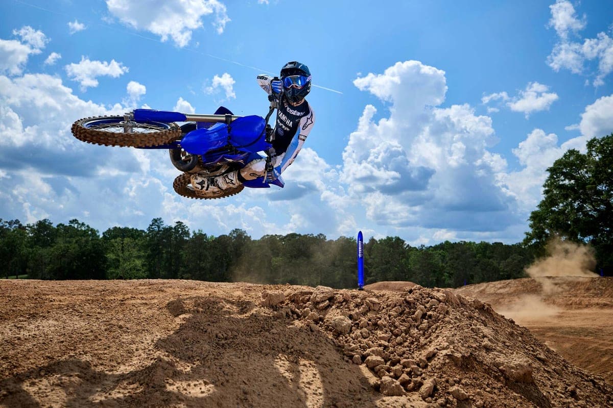 Rider in white and black gear scrubbing a blue Yamaha YZ250 sideways off a jump on a sandy motocross track under a cloudy sky