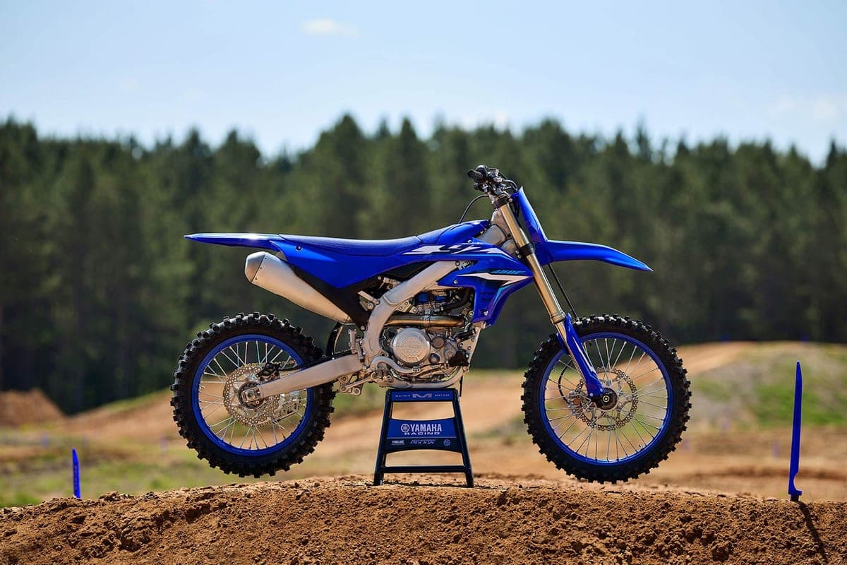 Yamaha YZ450F parked on a stand atop a dirt hill with pine trees in the background, side-on profile in soft daylight