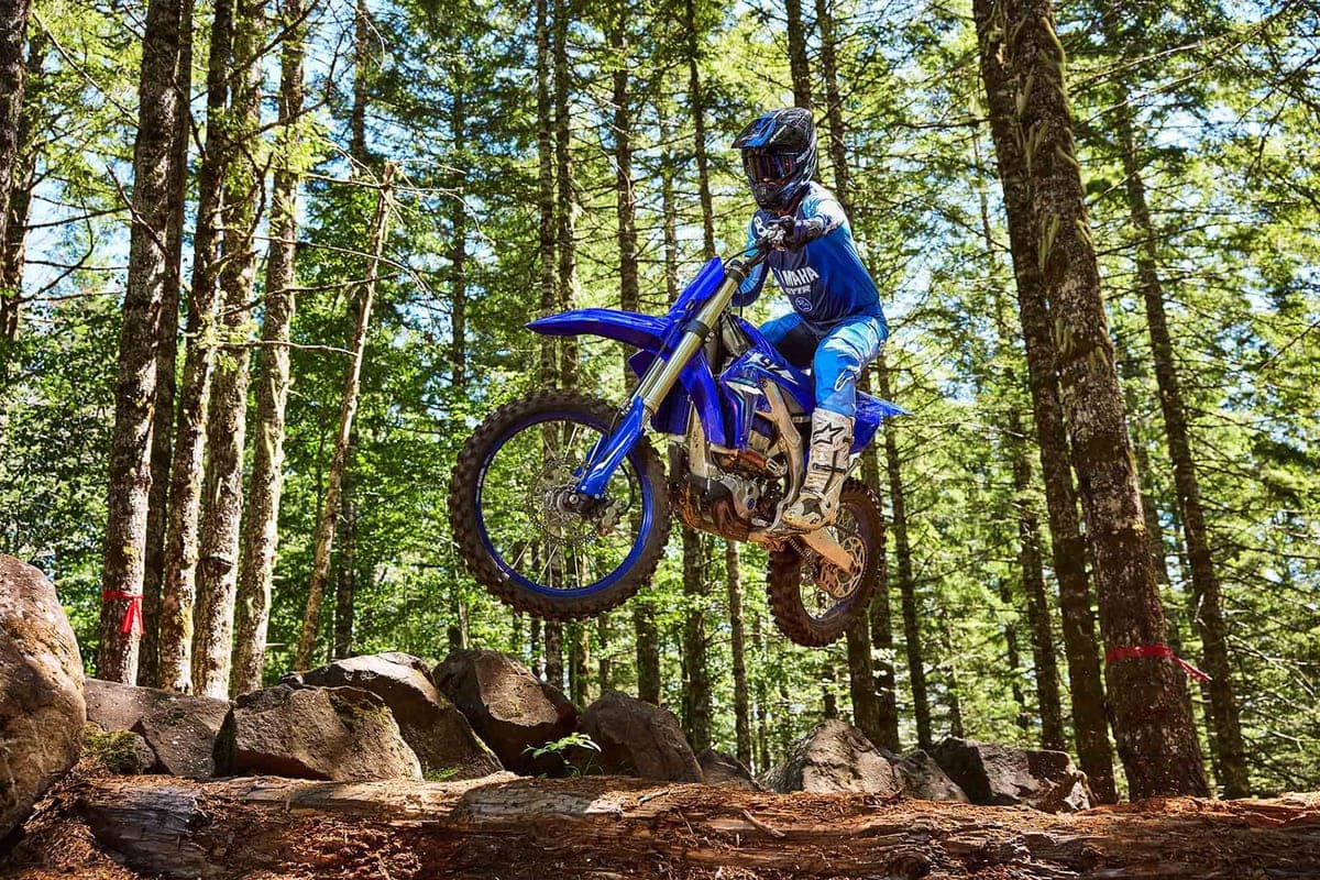 Rider in Yamaha blue gear leaping the YZ450FX off a rock ledge on a forested trail, front wheel tucked mid-air