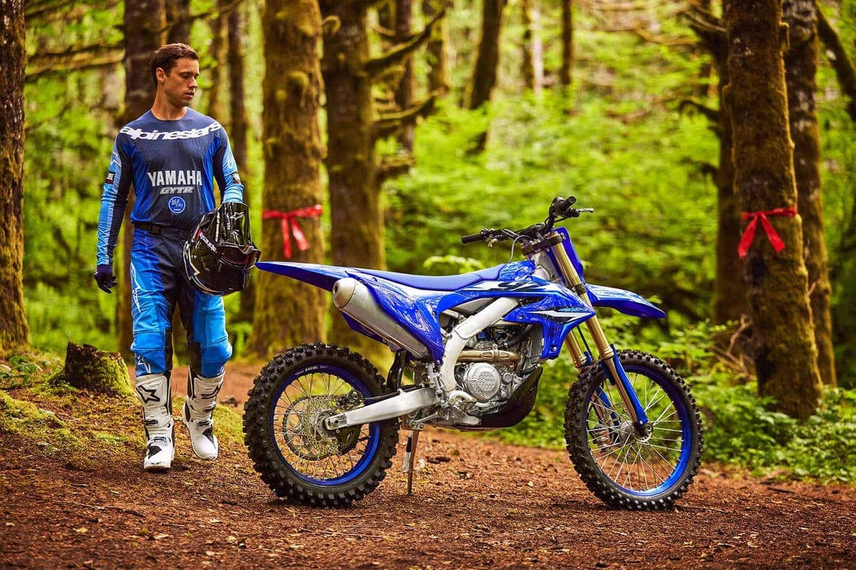 Yamaha GYTR rider walking beside a parked YZ450FX on a wooded dirt trail, showing the kickstand, side panel graphics and skid plate