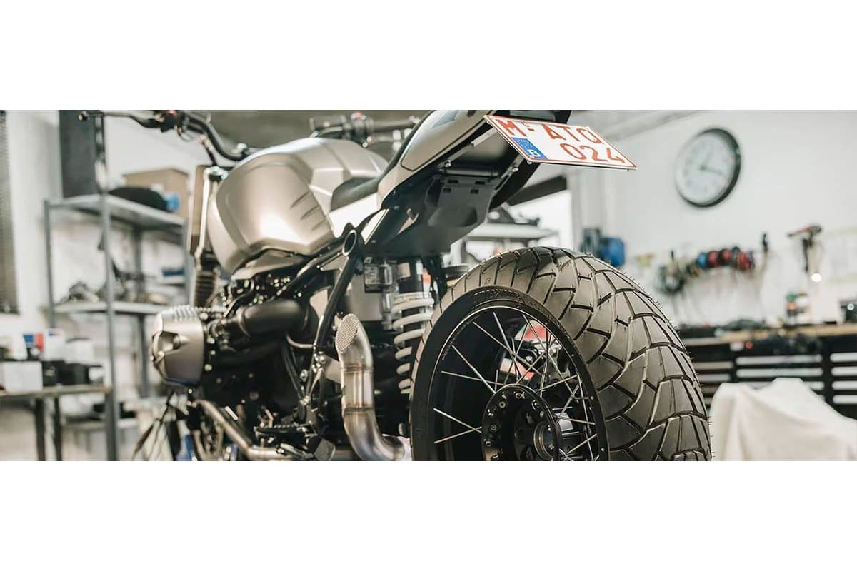 Bridgestone Battlax Adventurecross Scrambler AX41S: the tire mounted on the rear spoked wheel of a scrambler in a workshop, shot low from behind with the tread filling the frame