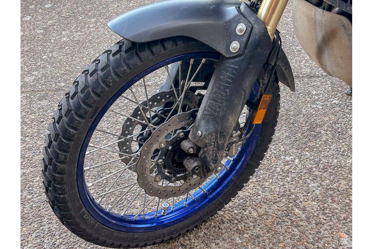 Heidenau K60 Scout: the front tire mounted on a blue-rimmed adventure bike wheel with the gold fork leg and brake disc in frame, shot on gravel