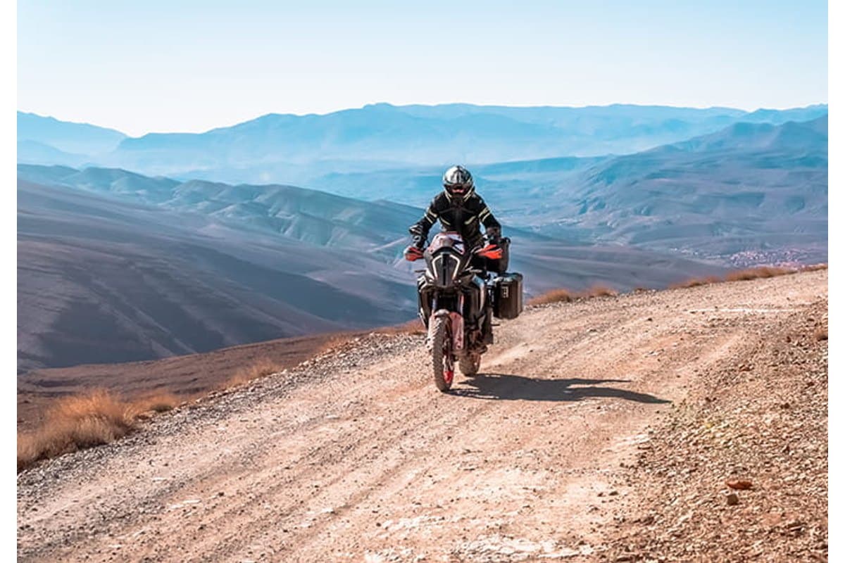 Mitas Enduro Trail: a rider climbing a gravel mountain road on a loaded adventure bike, arid ridgelines behind
