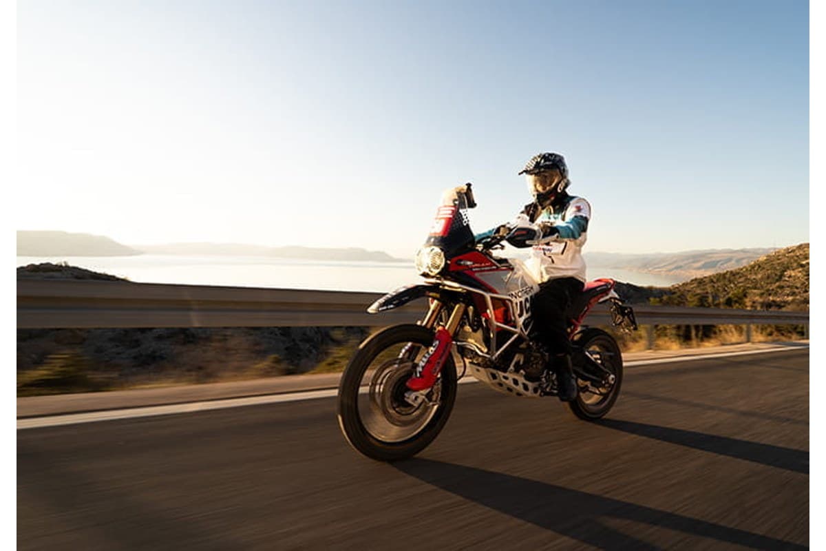 Mitas Enduro Trail-ADV 2: a rider on a red and white adventure bike leaning through a paved bend, a lake and hills behind