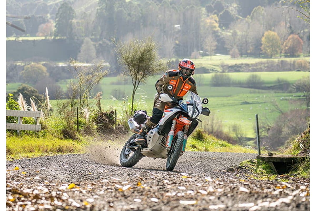 Mitas Enduro Trail+: a rider sliding a large adventure bike on a gravel track with dust off the rear, farmland behind
