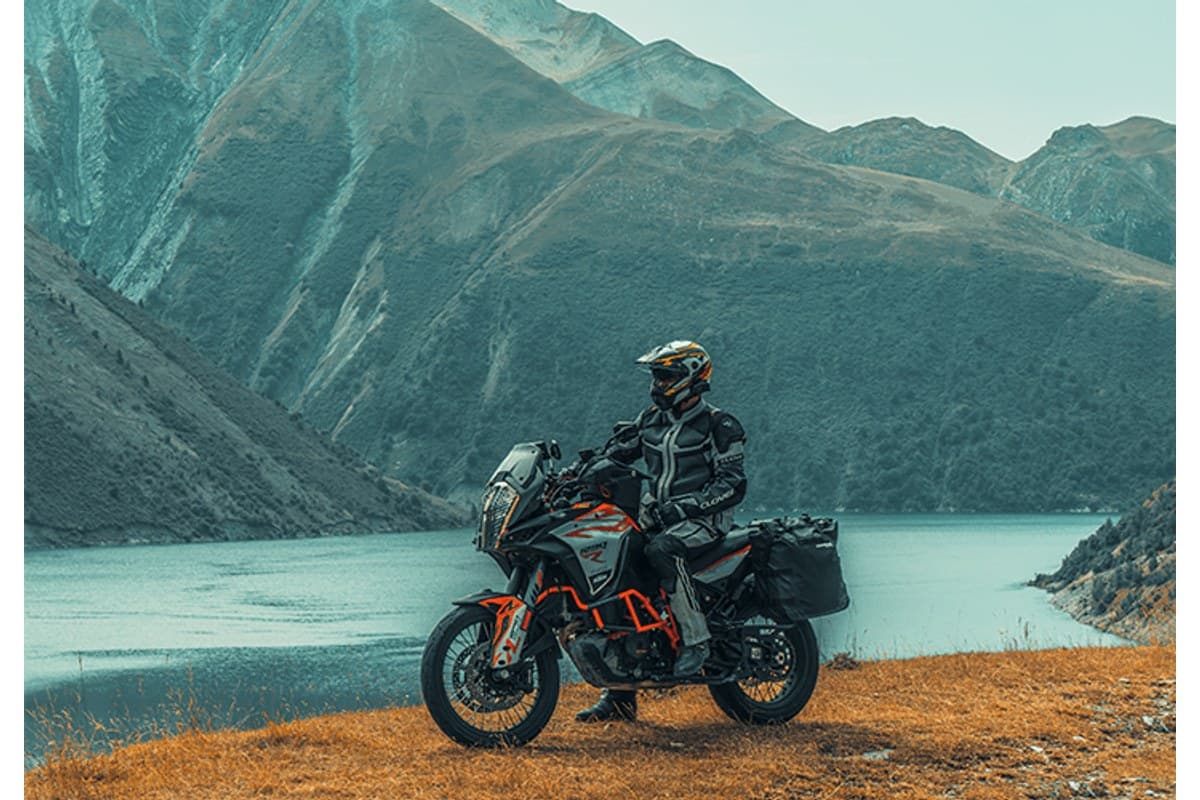 Mitas Enduro Trail-SP: a rider seated on an orange and black adventure bike with panniers, parked on a grassy bank above a mountain lake