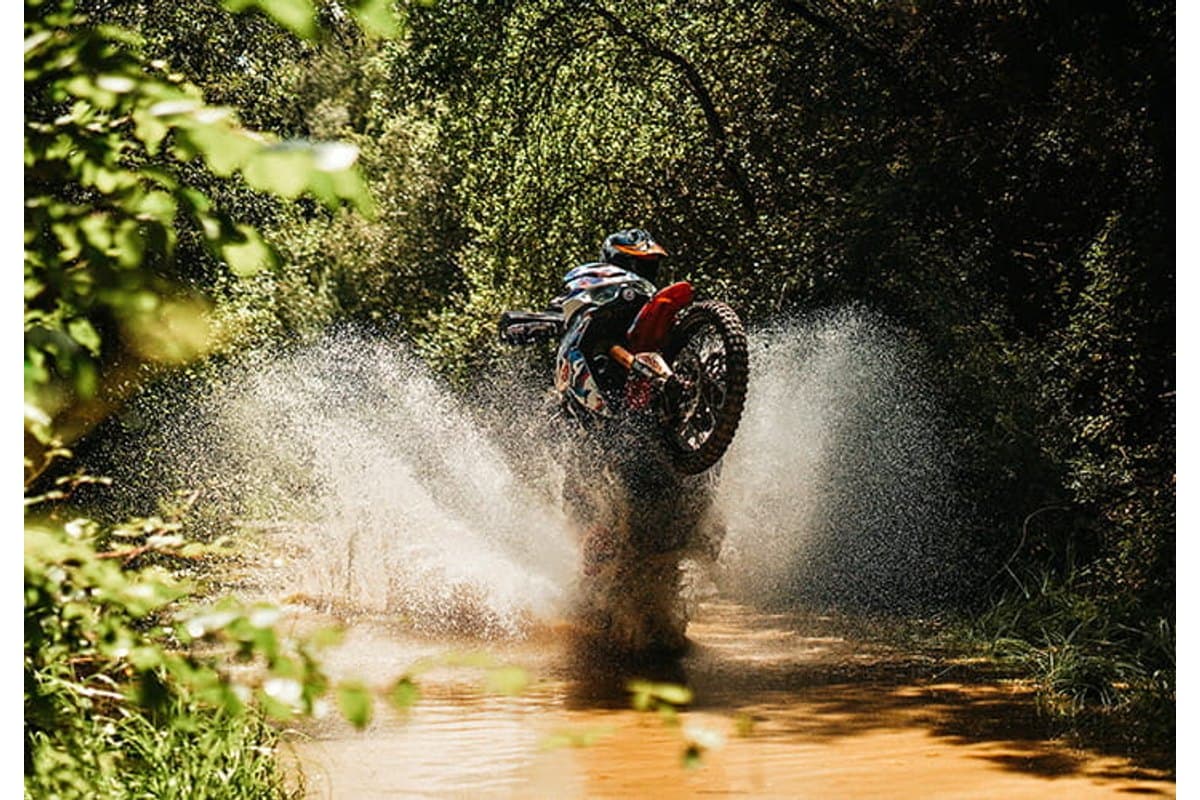 Mitas Enduro Trail-XT+: a rider lifting the front wheel through a muddy puddle on a wooded trail, knobby rear tire clearly visible