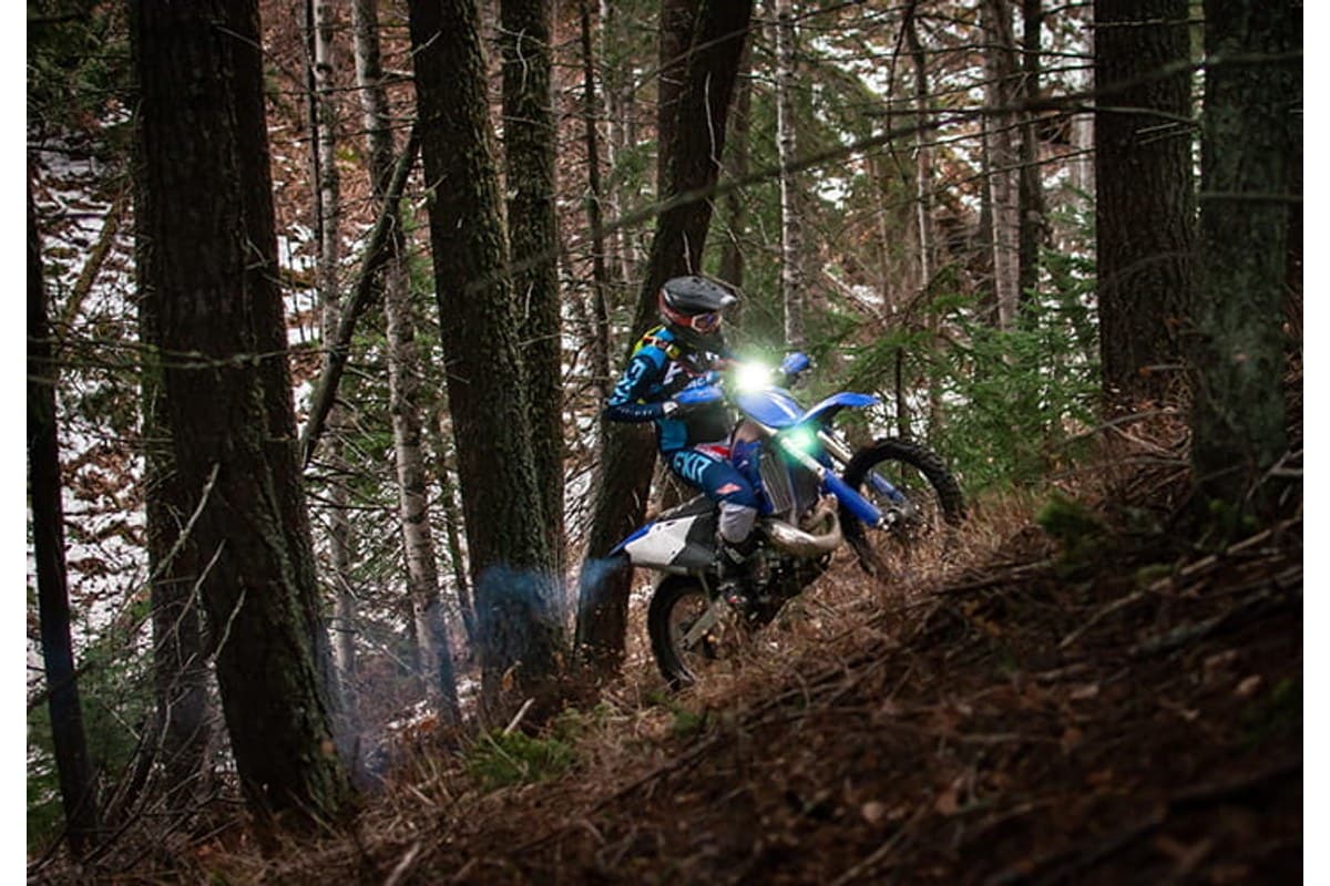 Mitas Stone King: a rider on a blue enduro bike, headlight on, climbing a steep bank through a dark pine forest