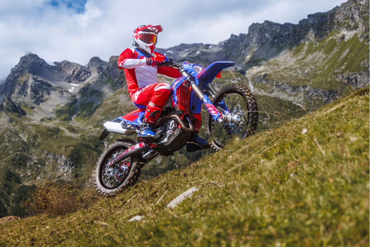 Rider in red and white gear powering the Beta 250 RR Race up a steep grassy alpine slope with rocky peaks behind