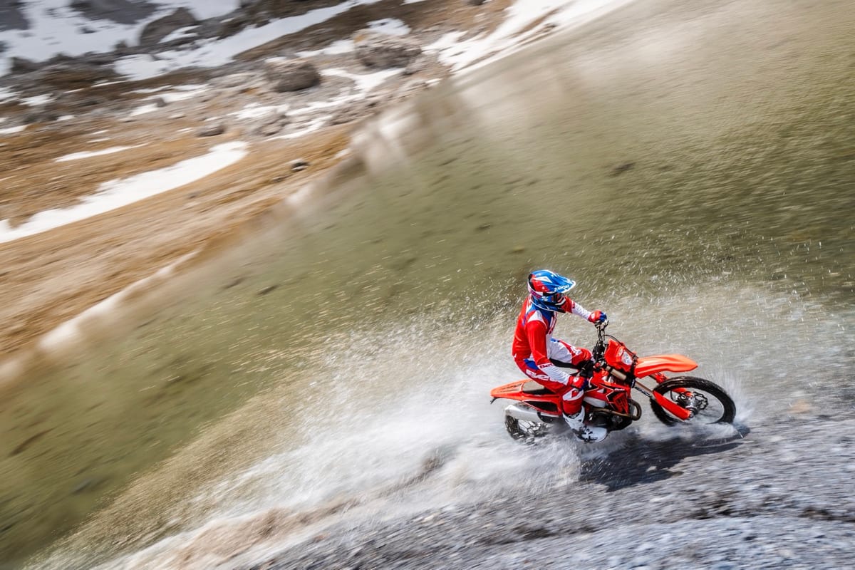 Rider in red, white and blue Beta gear railing a Beta 480 RR X-Pro through a rocky mountain stream, kicking up a wide spray of water