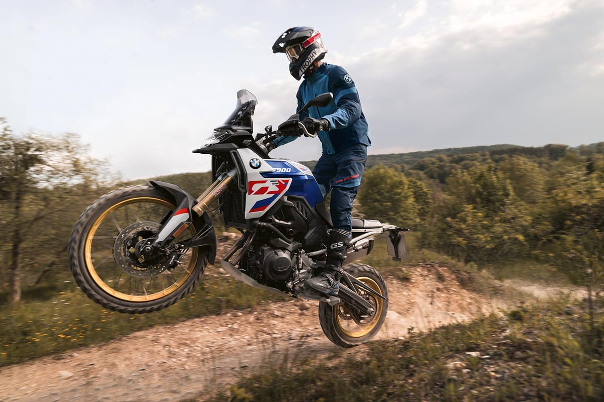 BMW F 900 GS in white, blue and red livery popping a wheelie on a dirt trail, rider in blue BMW GS gear standing on the pegs with gold-anodized fork and rims visible