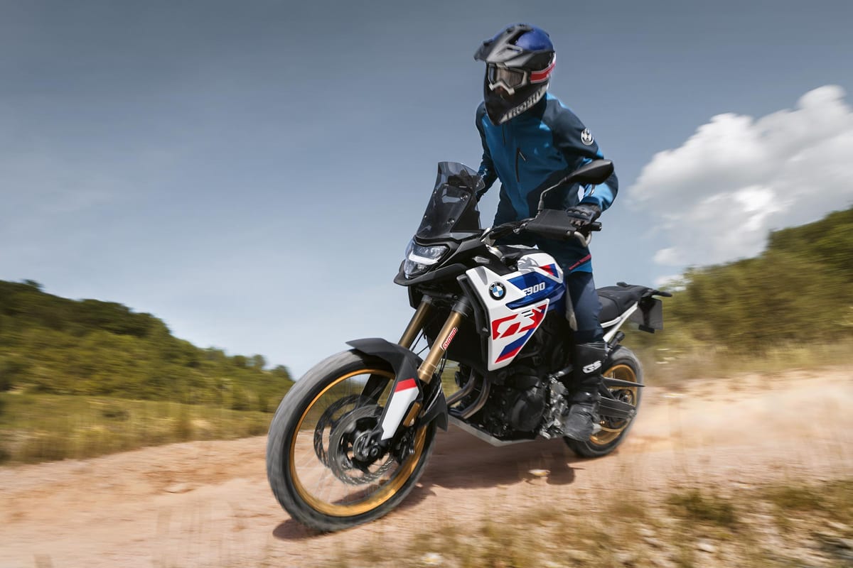BMW F 900 GS front three-quarter action shot leaning into a dusty dirt road corner, rider in blue jacket standing up, LED headlight and gold forks catching the light