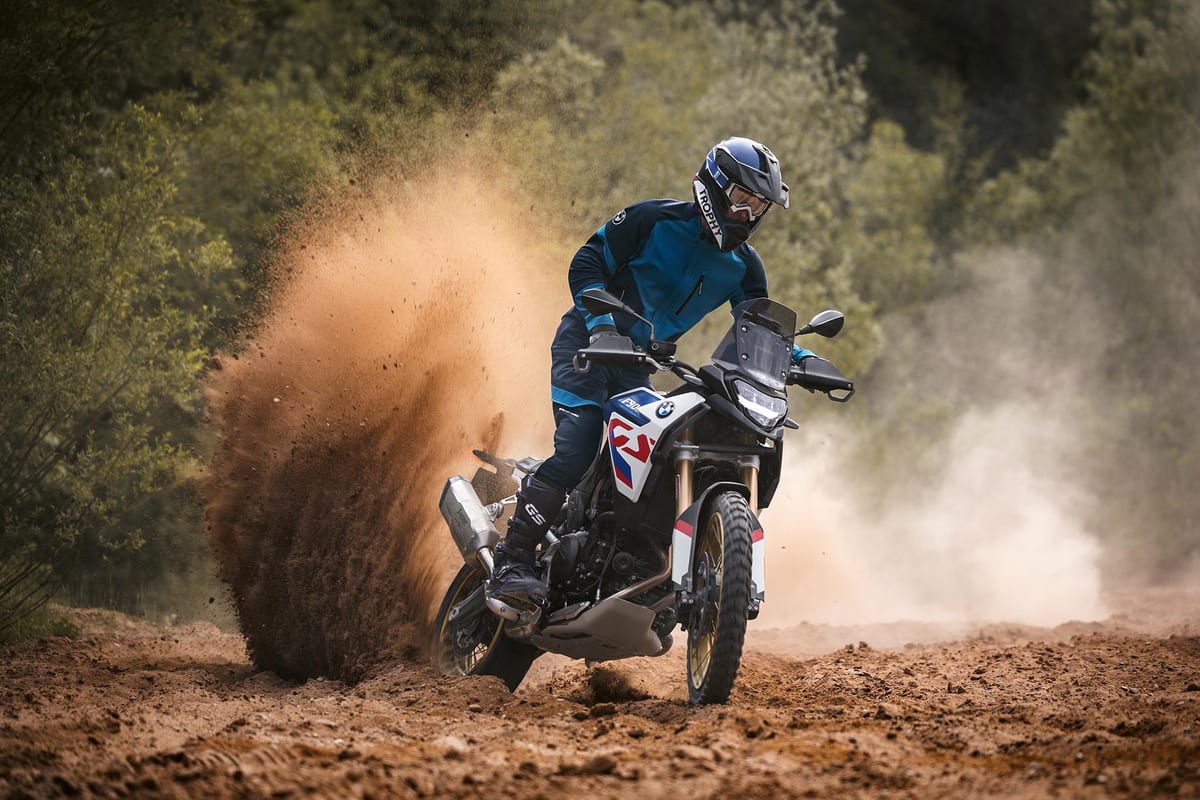 BMW F 900 GS kicking up a large dirt roost on a forest trail, rider in blue GS-branded gear seated with the twin exhaust cans and beak fender visible through the dust