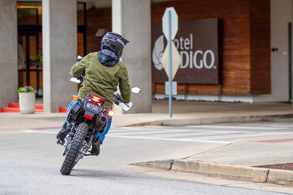 Rider in an olive jacket and blue jeans wheelies a white Honda XR650L away from camera on a city street outside a hotel entrance