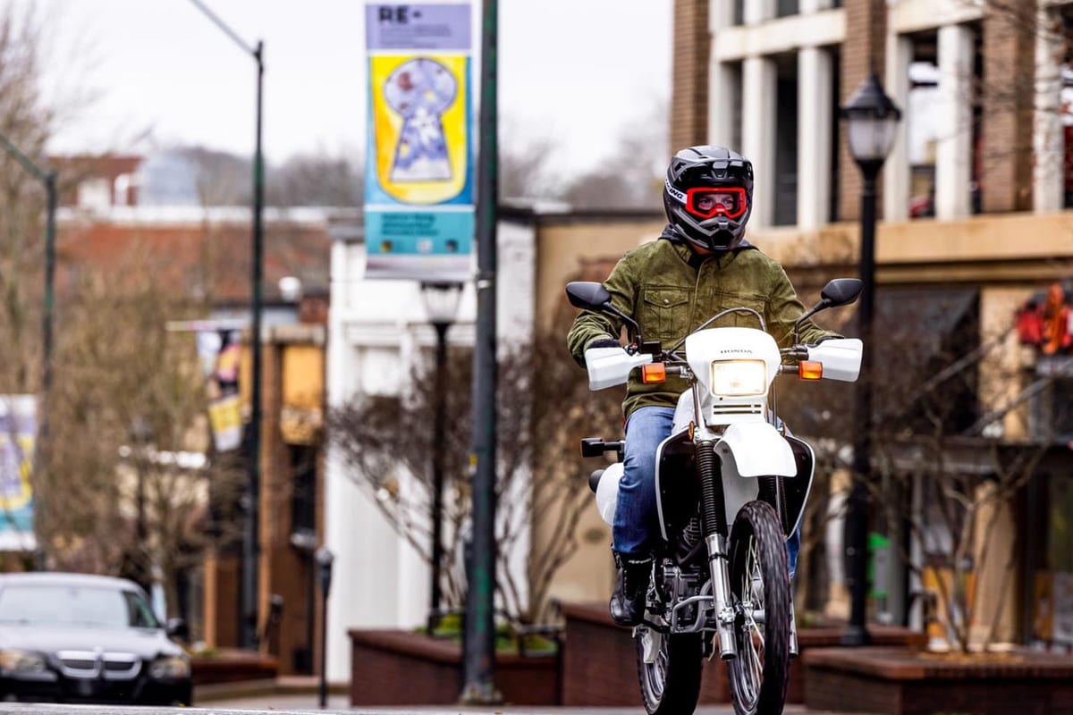 Rider in full-face helmet and goggles rides a white Honda XR650L toward camera down a downtown street past storefronts and a parked car