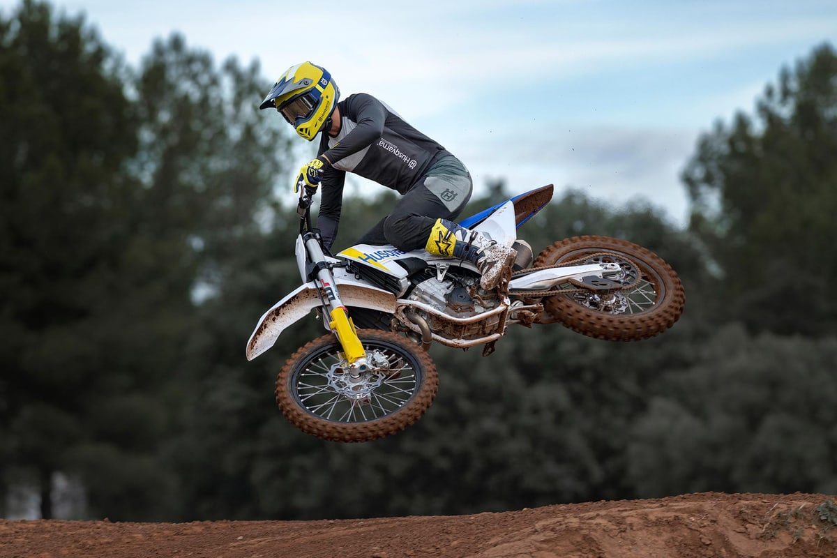 Rider in white and blue Husqvarna gear whipping the FC 450 over a jump against a treeline