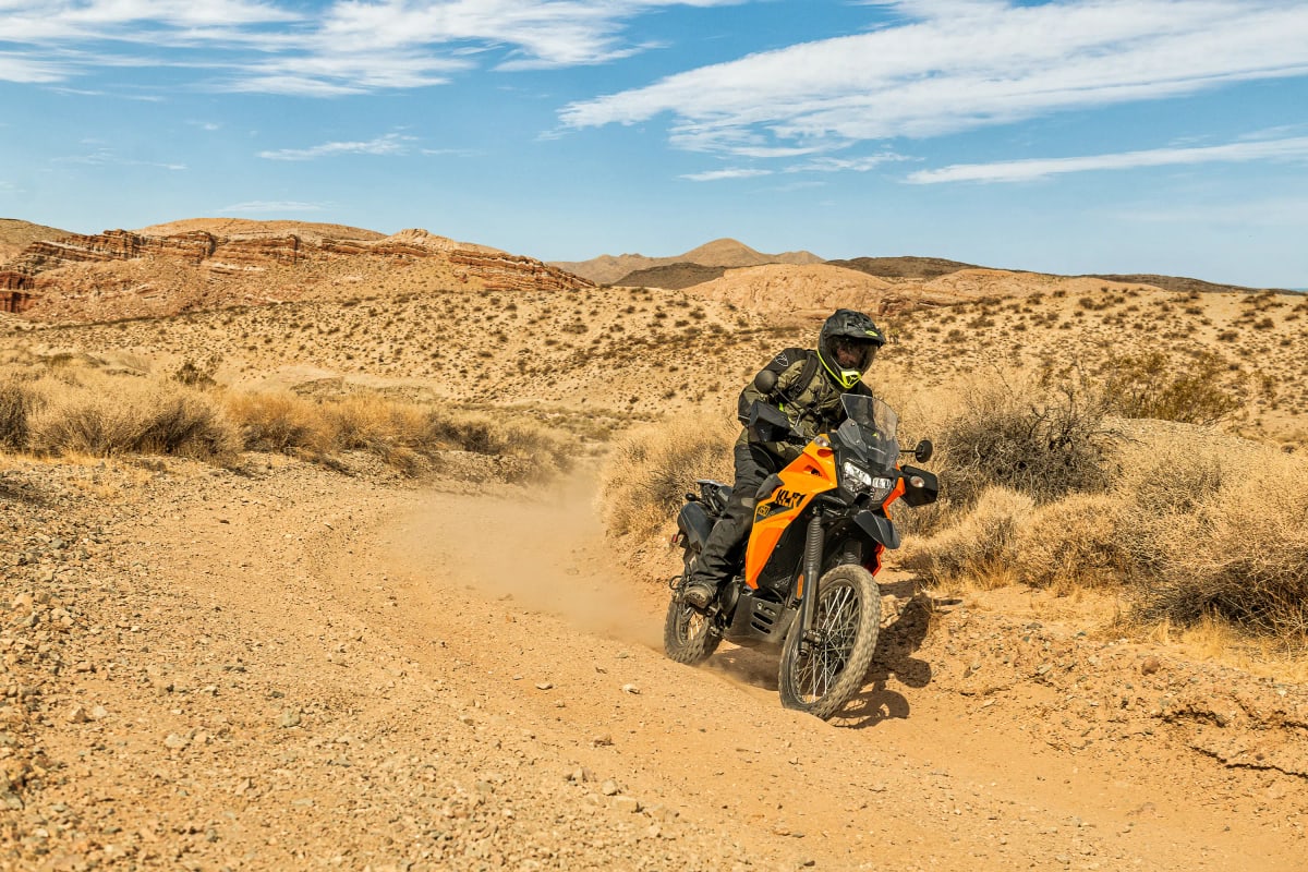 Rider on a yellow Kawasaki KLR650 S on a sandy desert trail with red rock behind