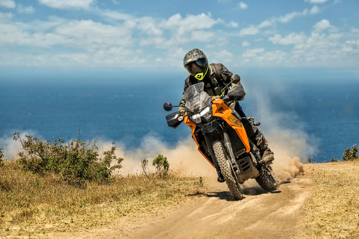 Rider sliding a yellow Kawasaki KLR650 S through dust on a coastal dirt road above the sea