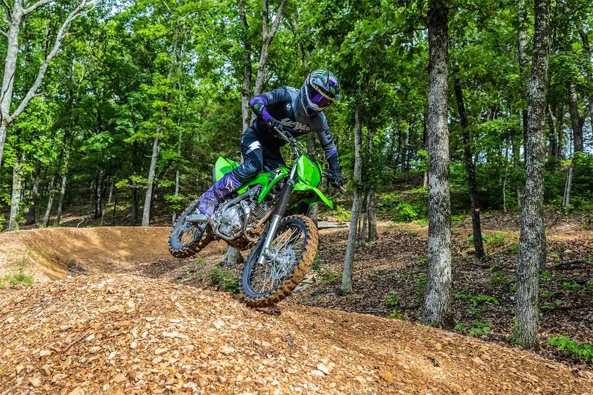 Rider in dark gear jumping a Lime Green Kawasaki KLX230R S over a wooded trail rise