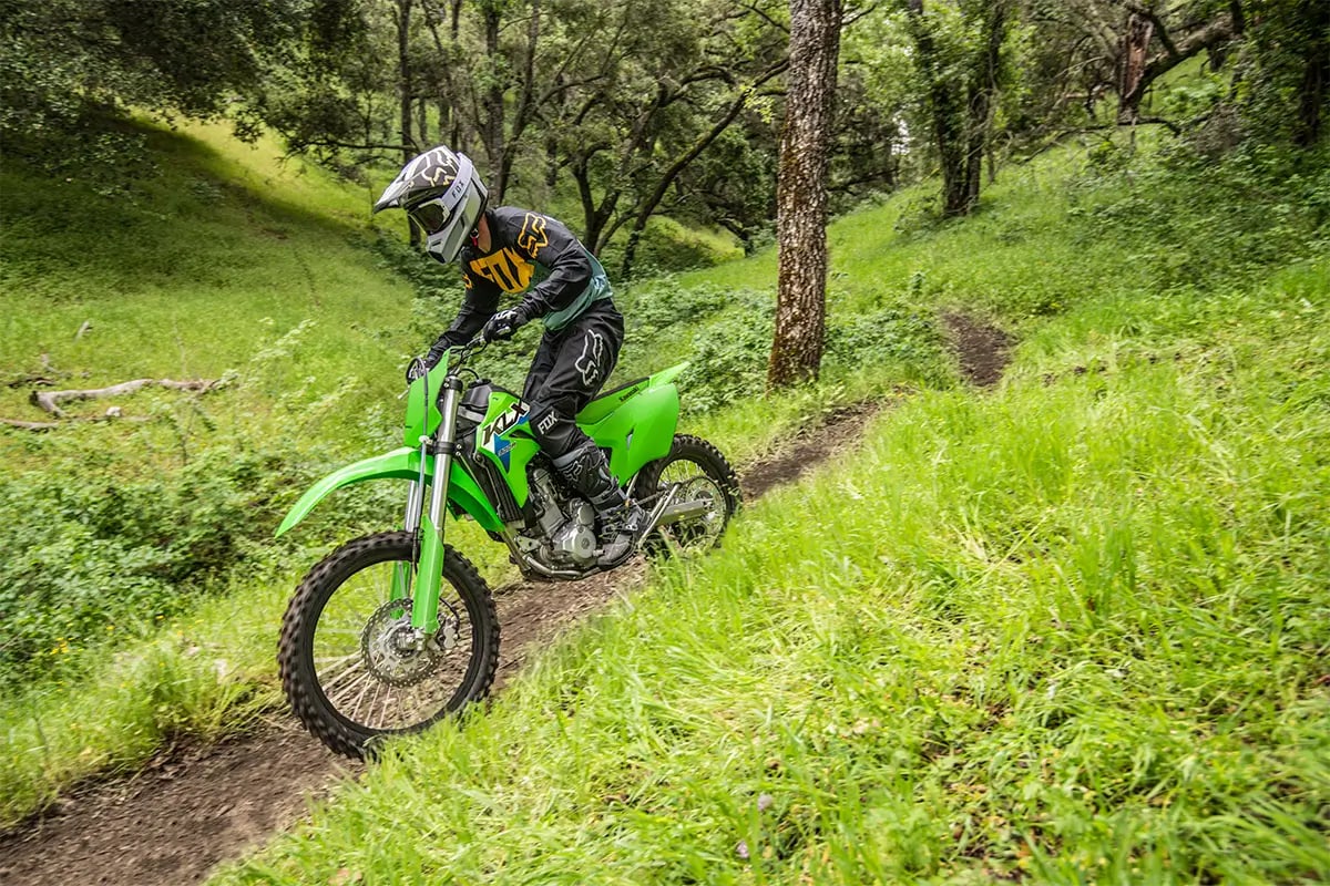 Rider in grey and yellow gear climbing a narrow wooded singletrack trail on a Lime Green Kawasaki KLX300R