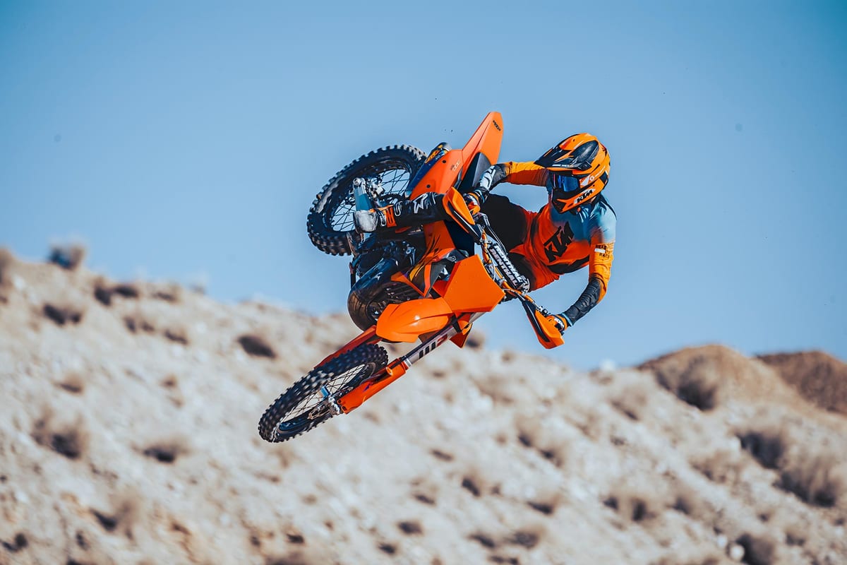 Rider in orange and blue KTM gear whipping a 300 SX sideways off a jump against a clear blue sky on a rocky hillside track