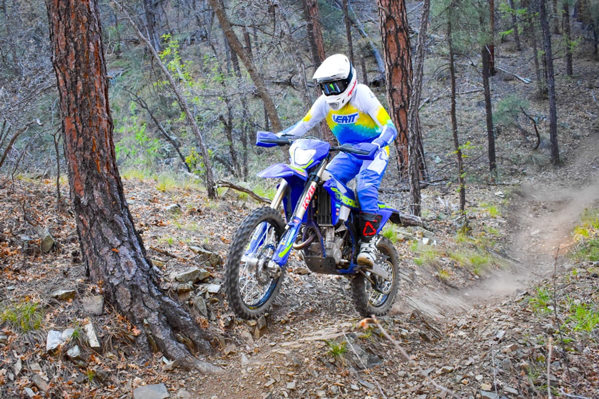 Rider in white and yellow Leatt gear powering the blue Sherco 250 SEF Factory up a rocky forest trail, rear wheel kicking up dust