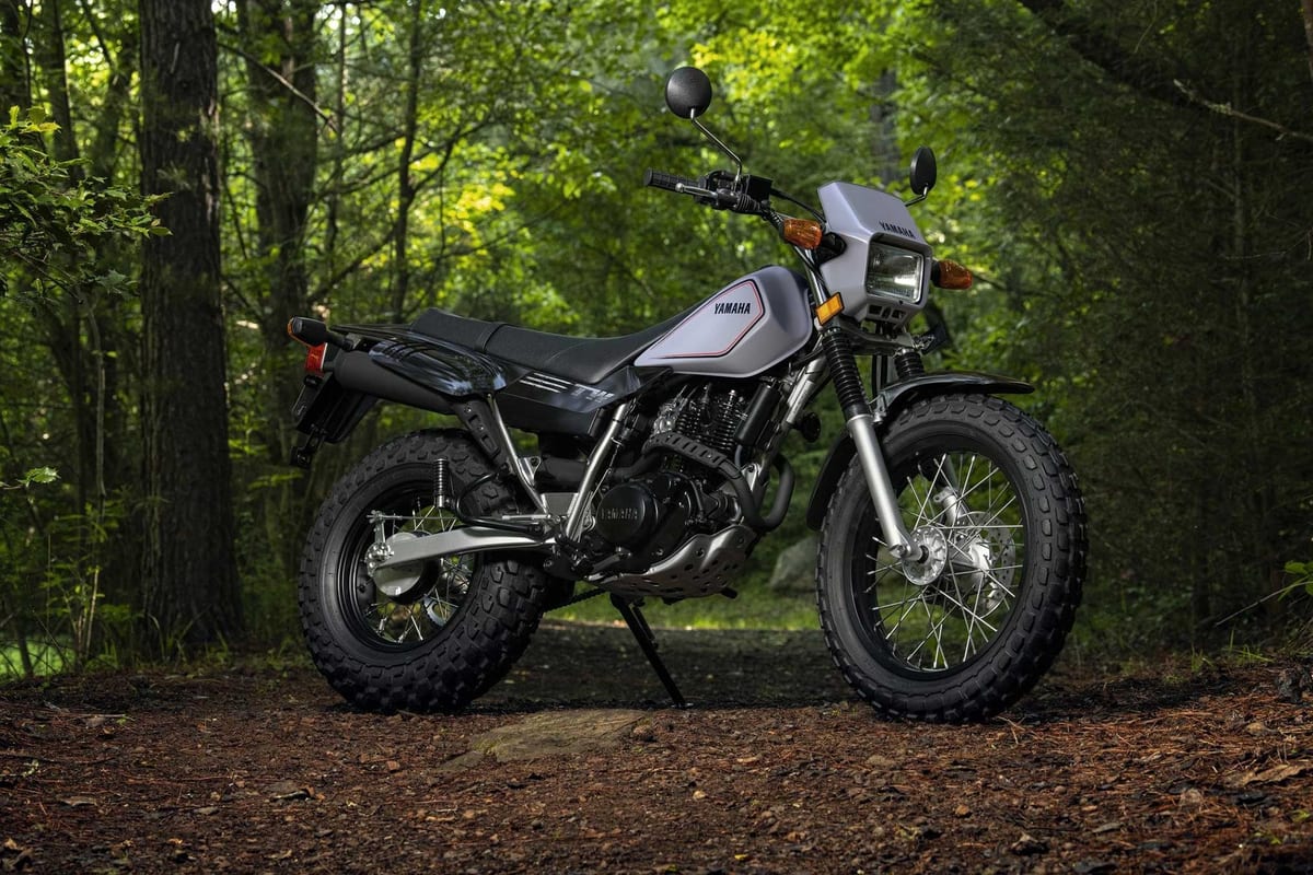 Yamaha TW200 in silver, angled studio shot in a forest clearing highlighting the fat knobby tires, spoked wheels and stubby exhaust