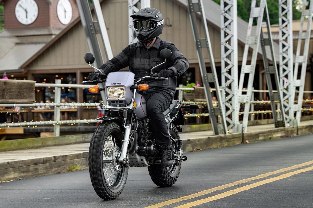 Rider in dark plaid jacket and full-face helmet riding a silver Yamaha TW200 across a small-town bridge with string lights and a clock tower behind