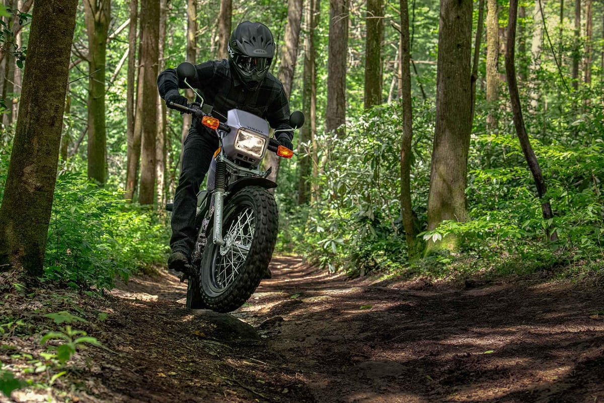 Rider in black gear popping the front wheel of a silver Yamaha TW200 on a shaded dirt trail, knobby tire kicking up loose soil