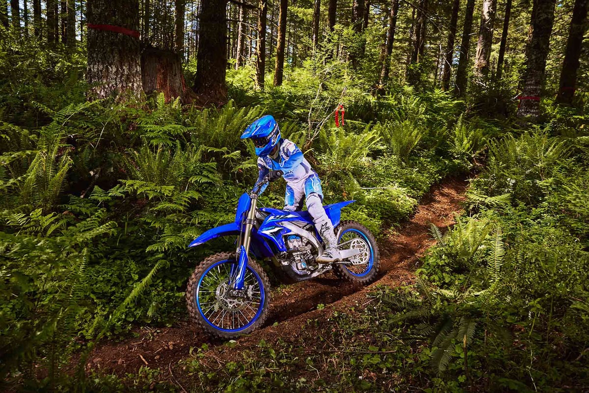 Rider in blue and white gear leaning the YZ250FX down a shaded forest trail edged with ferns and course-marker tape