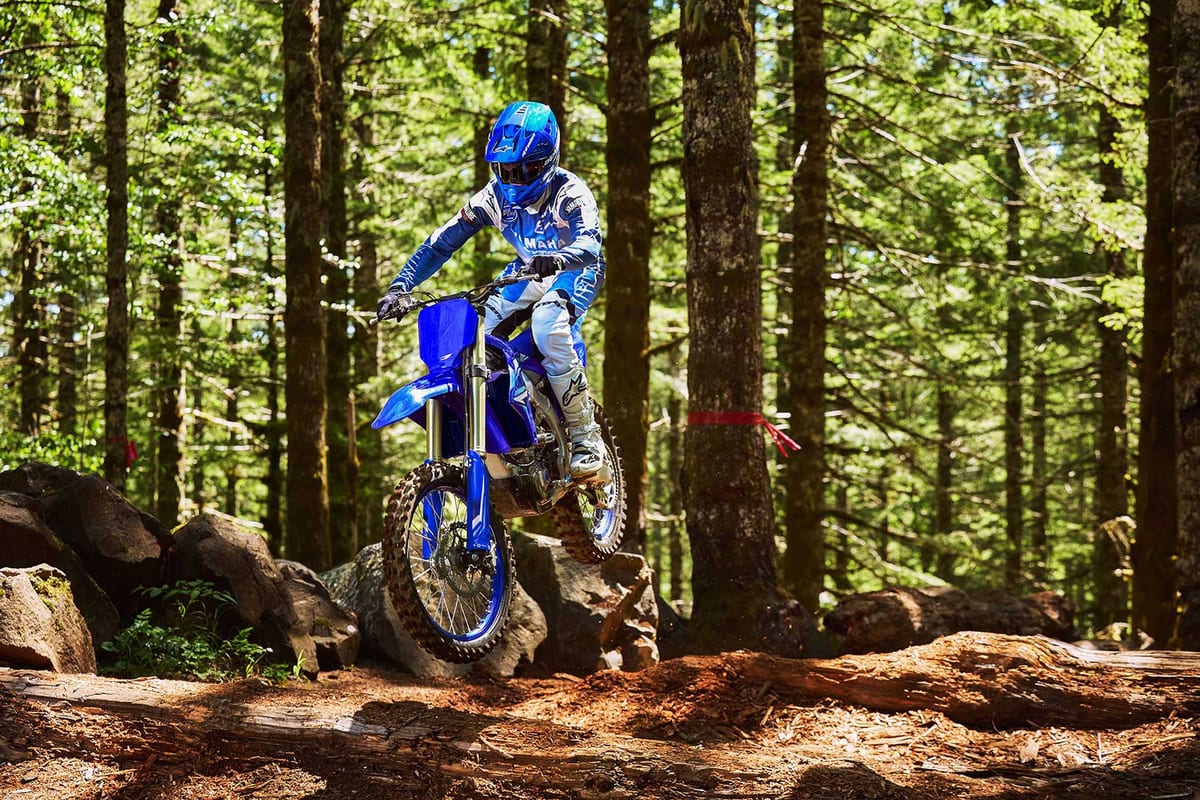 Yamaha YZ250FX airborne off a rock ledge on a wooded trail, rider standing on the pegs amid tall pines